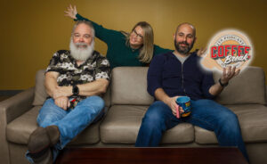 Podcast team sitting on the couch smiling with one member standing behind the couch with their arms stretched out.